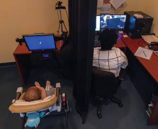 Person works at computer, baby in bouncer on floor separated by a dark curtain.