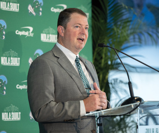 Man in suit speaks at podium, hands clasped, against a green "NOLA BUILT" banner.