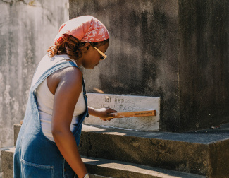 Photograph: Woman in overalls presses a brush onto a stone tomb in a cemetery.