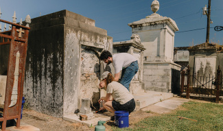 Two people cleaning an old, weathered tomb in a sunny cemetery.