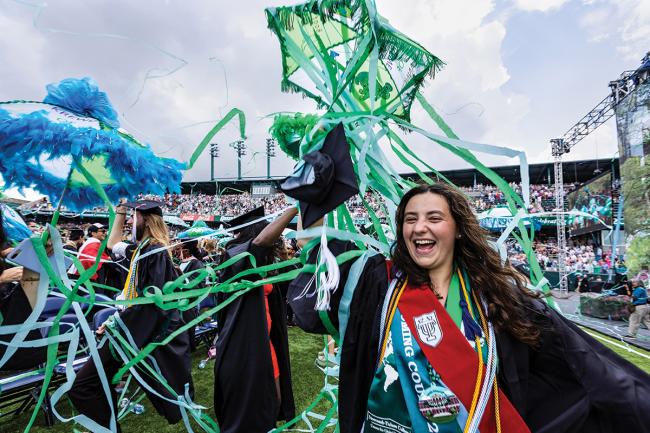 Crowd of graduates celebrate as streamers come down on a stadium field. A graduate in the foreground holds decorated umbrella.
