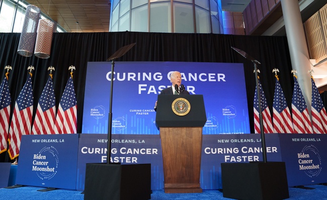 President Biden speaks from stage with signs that read Curing Cancer Faster 