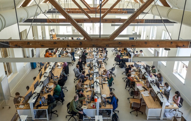 Large open-plan workspace with high ceilings and wooden beams, filled with students working at long shared desks with computers.