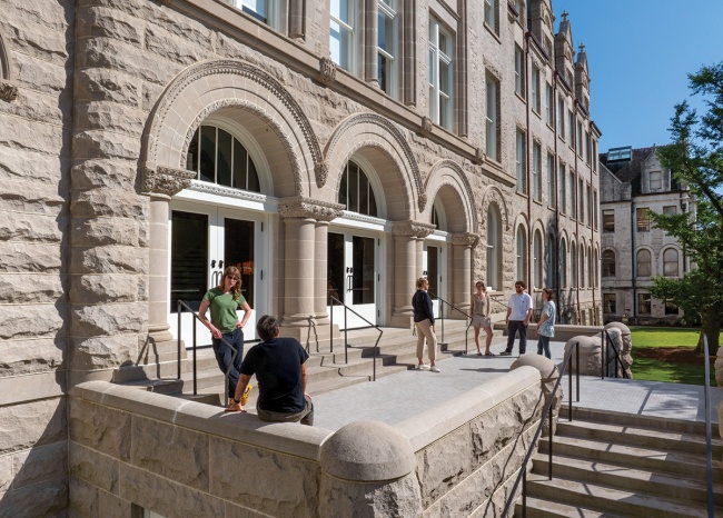 Several people gather outside a historic stone building on Tulane University's campus, chatting in small groups. The building features arched doorways, decorative stonework, and tall windows, with a bright blue sky and green lawn in the background.