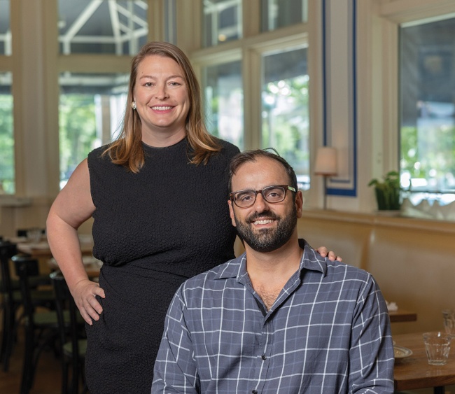 Restaurant owners Alon and Emily Shaya pose together inside a bright, stylish dining space, Saba. Emily, wearing a sleeveless black dress, stands with one hand on her hip and the other resting on Alon’s shoulder. Alon, seated and wearing glasses and a blue plaid shirt, smiles toward the camera. Large windows and tables are visible in the background.