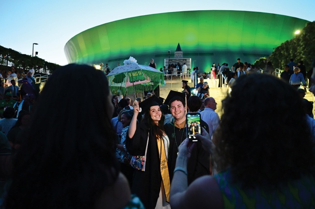 graduates in cap and gown pose for a photo outside a green-lit Superdome at dusk, surrounded by a crowd of people