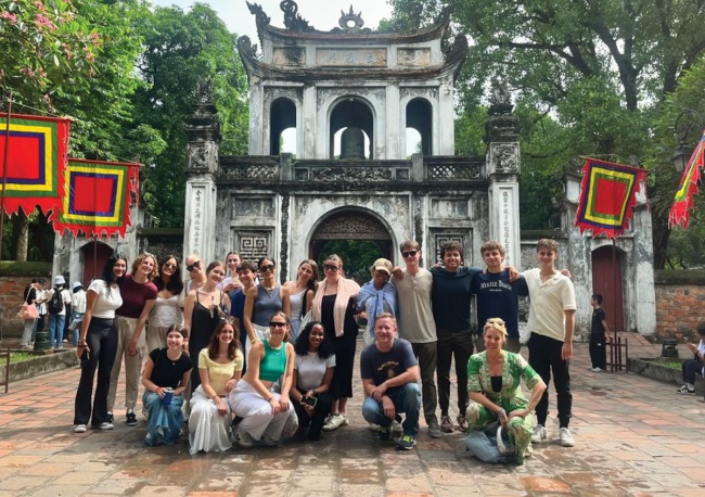 A group of Altman Scholars poses in front of an ancient stone gate with three arched openings. Colorful flags hang on either side, and lush green trees surround the scene. 