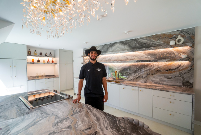A smiling man in a cowboy hat stands in a modern kitchen with marble countertops and backsplash. Soft lighting creates a warm, inviting atmosphere.