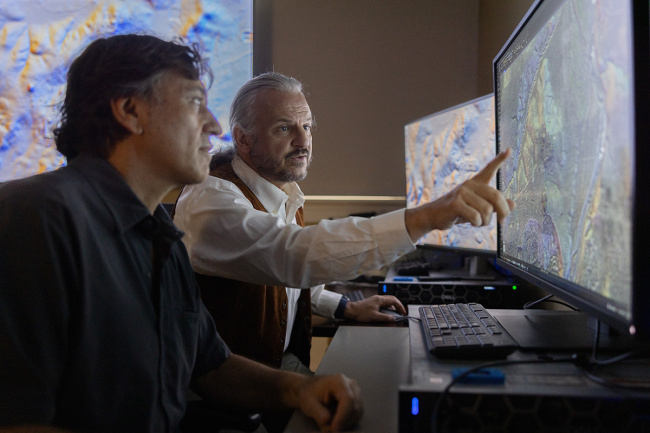 Two men analyze a detailed map on a large monitor. One points at the screen, focusing intently. The room is dimly lit, suggesting concentration.