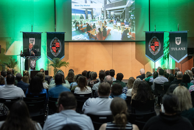 A speaker addresses an audience at a conference, standing at a podium with Tulane banners. A large screen displays an image of a market scene. 