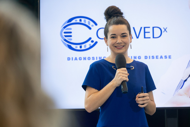 Smiling woman in blue dress holding a microphone, presenting in front of a screen with logo. 