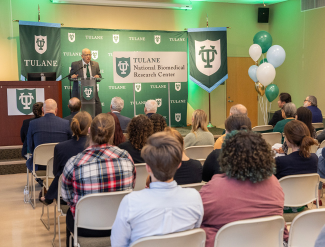 A speaker at a podium addresses an audience. Behind are banners and a sign for the Tulane National Biomedical Research Center.