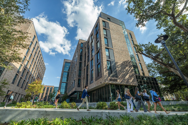 A group of students walks past modern, multi-story brick buildings under a blue sky with scattered clouds. The scene is lively and academic.