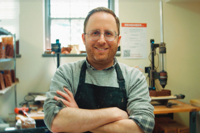 Smiling person with glasses and beard, arms crossed, wearing a black apron in a workshop. Shelves and tools create a cozy, industrious atmosphere.