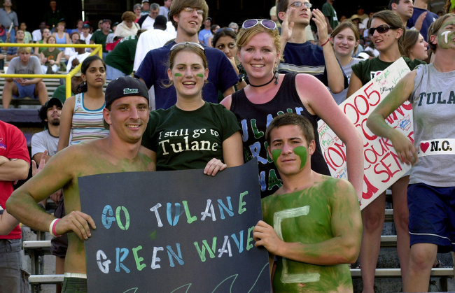 Fans in a stadium cheer energetically, holding a sign that reads "Go Tulane Green Wave." Faces are painted green, expressing school spirit and excitement.